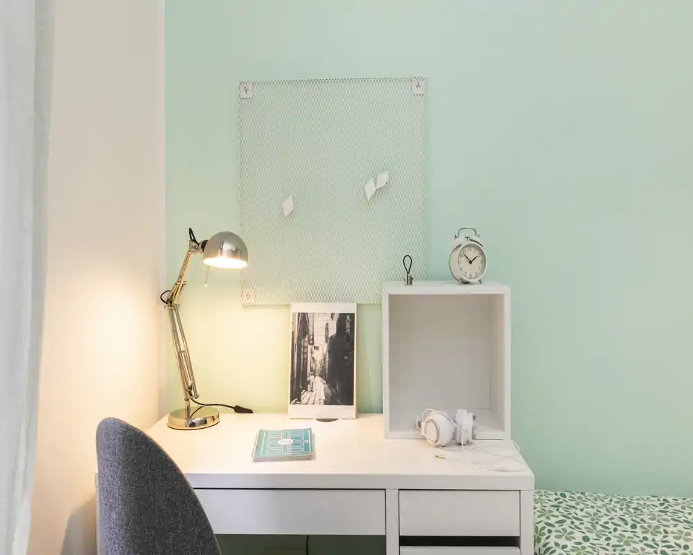 Rincón de dormitorio ordenado con escritorio blanco, lámpara de trabajo y un pequeño borde de la cama visible; pared verde menta y accesorios sencillos que aportan un aspecto moderno y tranquilo.
