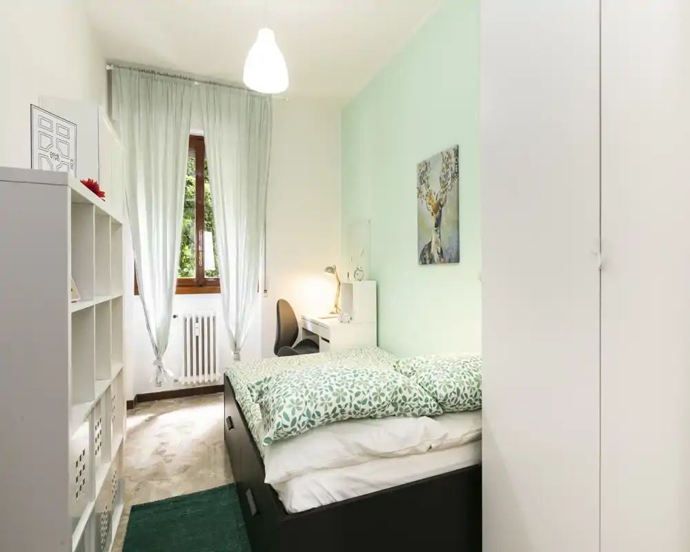 Dormitorio luminoso y ordenado con cama doble con ropa de cama verde estampada, un escritorio compacto junto a la ventana y un cuadro decorativo — ideal para una persona o pareja.