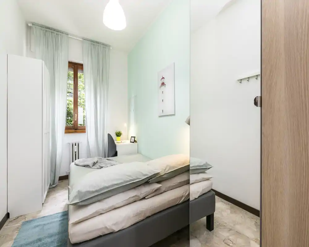 Camera singola luminosa e ben allestita con letto comodo, comodino e finestra che lascia entrare luce naturale. Parete accentata color menta e arredi semplici creano un'atmosfera tranquilla e accogliente.