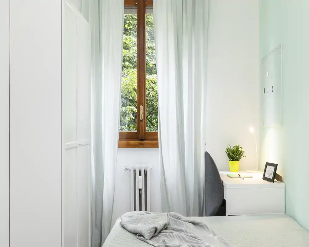 Camera singola luminosa e minimal con letto ben rifatto, tende leggere e una scrivania vicino alla finestra. La luce naturale e le tonalità pastello creano un’atmosfera fresca e accogliente, perfetta per affitti brevi.