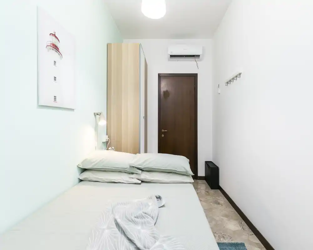 Camera da letto compatta e luminosa con letto matrimoniale ben rifatto, lampada da comodino e armadio vicino all'ingresso. Parete color menta e pavimento in marmo donano un aspetto moderno e pulito.
