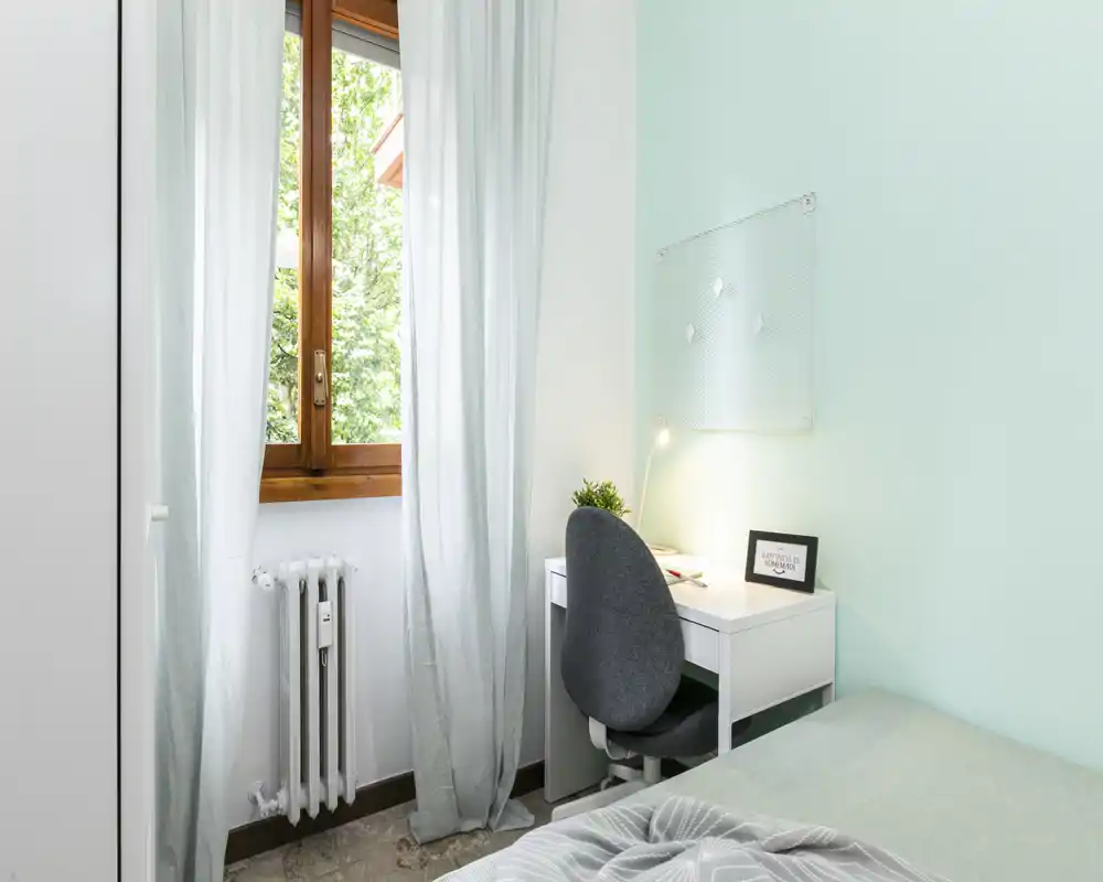 Camera luminosa e compatta con letto singolo, scrivania e sedia accanto a una finestra soleggiata — ideale per studio o soggiorni brevi.