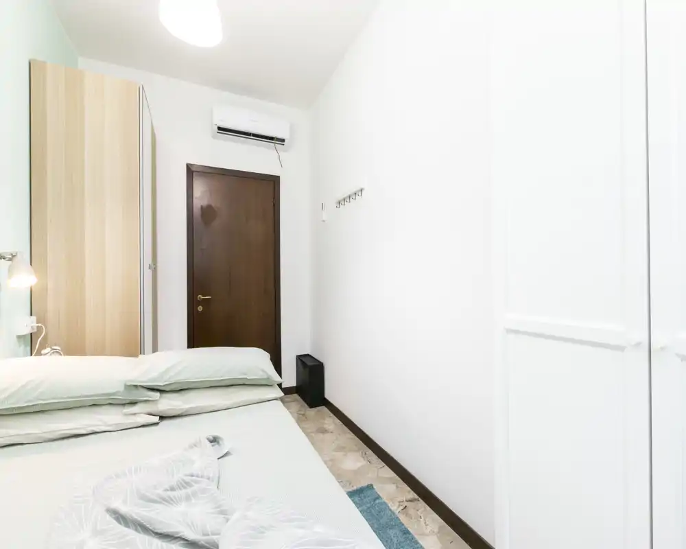 Camera da letto compatta e luminosa con letto matrimoniale sistemato, armadio e condizionatore. Lenzuola chiare e tonalità neutre creano un ambiente pulito e rilassante, perfetto per soggiorni brevi.