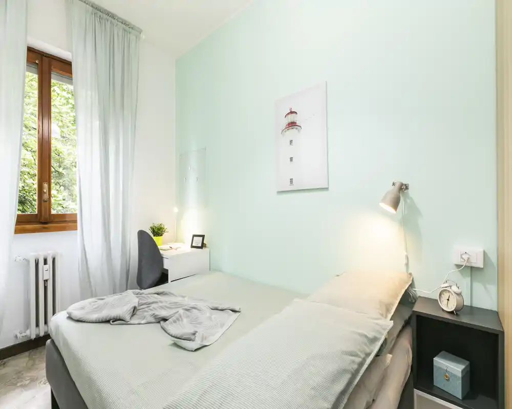 Camera da letto luminosa in tonalità pastello con letto ben rifatto, lampada da comodino e una piccola scrivania vicino alla finestra — ariosa e ben allestita per gli annunci.