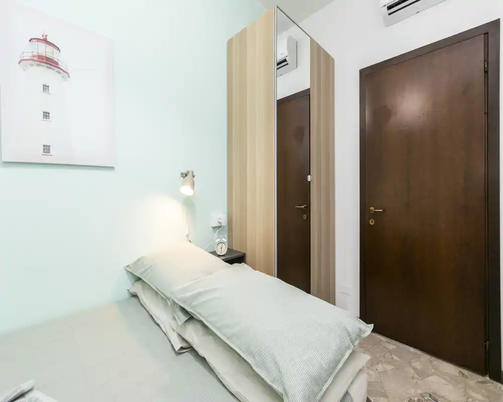 Camera da letto piccola e ordinata con letto singolo, lampada da comodino e armadio con specchio. Parete color menta e biancheria curata creano un ambiente rilassante, perfetto per soggiorni brevi.