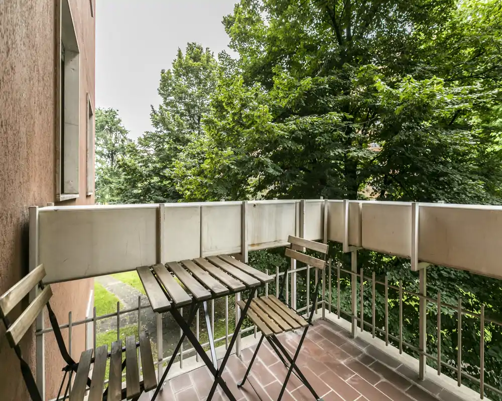 Piccolo balcone arredato con tavolino in legno e sedie pieghevoli, affacciato su alberi rigogliosi — ideale per colazione o momenti di relax all'aperto.