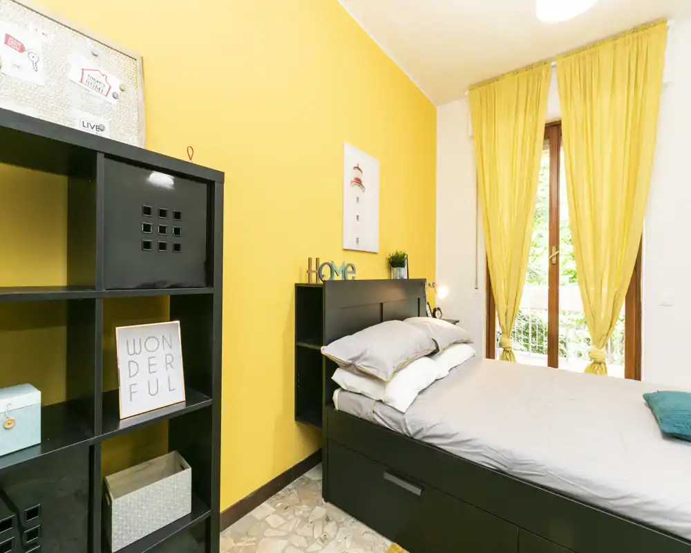 Camera da letto luminosa e accogliente con letto matrimoniale ben rifatto, parete gialla d'accento e ampia finestra con tende abbinate che lasciano entrare luce naturale.