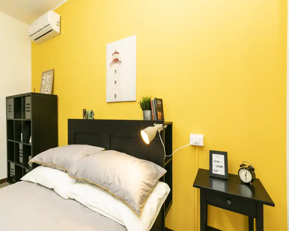 Camera da letto moderna e luminosa con letto matrimoniale, comodino, lampada e dettagli decorativi su una parete gialla vivace.