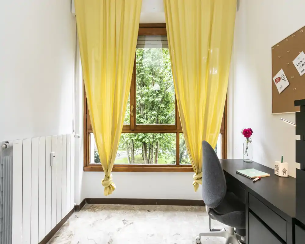 Angolo di camera luminoso con ampia finestra incorniciata da tende gialle, una scrivania compatta e una sedia — luce naturale intensa e arredo minimale e accogliente.