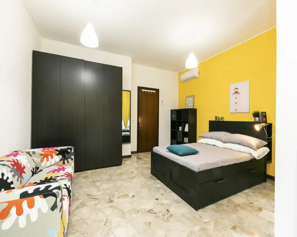 Ampia camera da letto luminosa con letto moderno nero con cassetti, grande armadio e una poltrona colorata davanti a una parete accentuata di colore giallo.