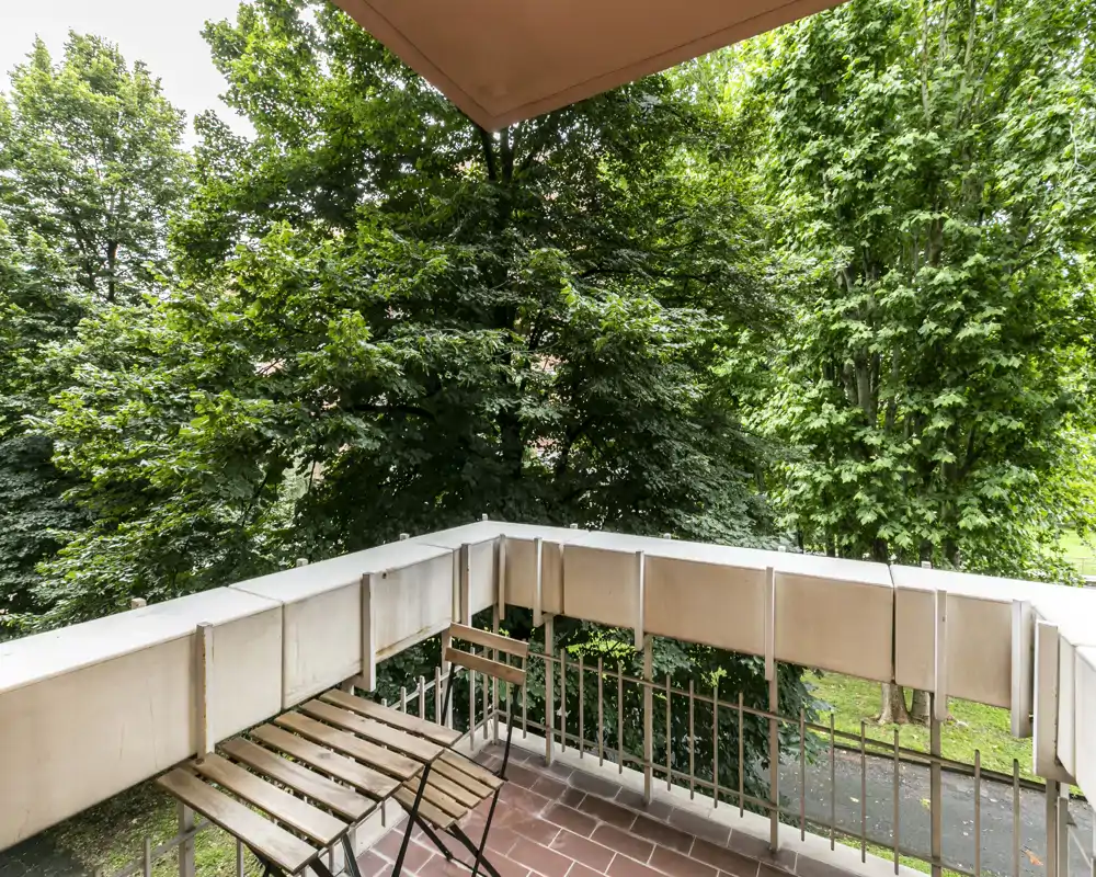 Balcone soleggiato con un piccolo tavolo bistro in legno e sedie pieghevoli, affacciato su alberi rigogliosi — angolo esterno tranquillo per il caffè del mattino.