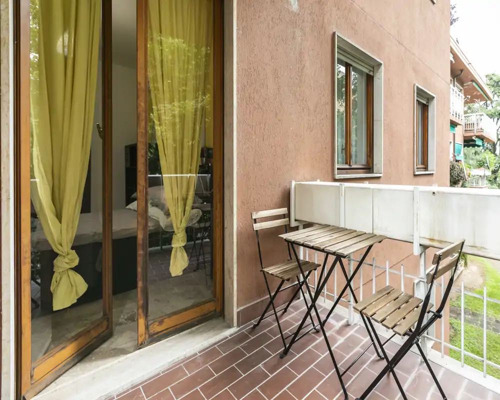 Piccolo balcone affacciato sul giardino con un tavolino bistro in legno e due sedie; attraverso le porte finestra si intravede una camera da letto con tende gialle e letto parzialmente visibile.