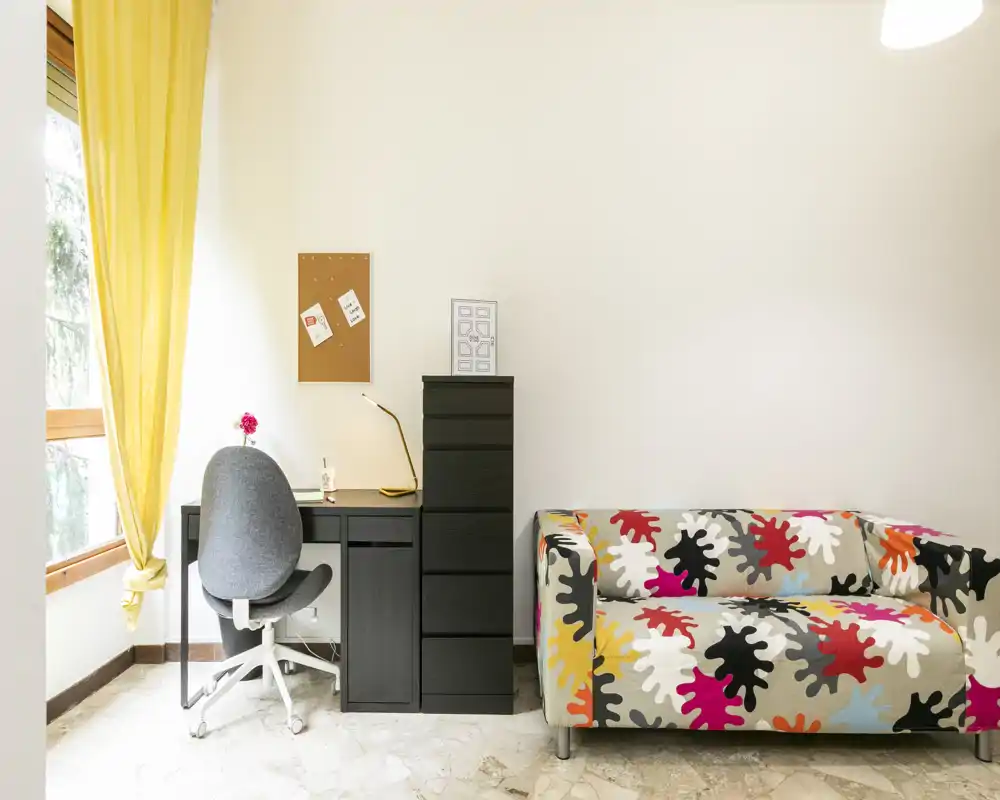 Zona giorno luminosa e moderna con divano colorato, angolo studio compatto e ampia finestra — ideale per un confortevole monolocale.
