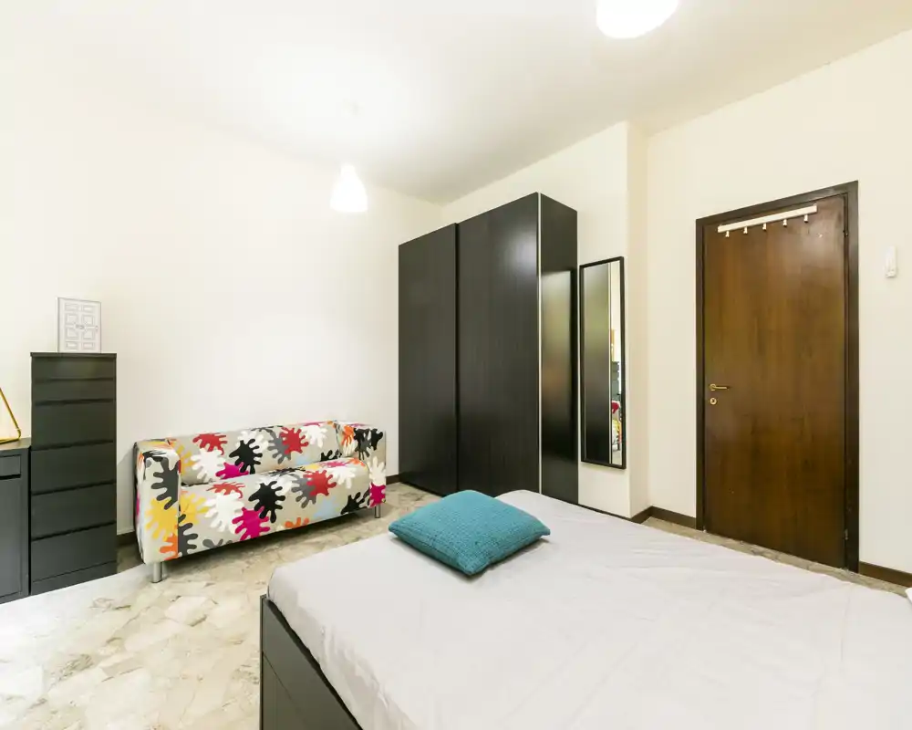Camera da letto luminosa e ordinata con letto matrimoniale visibile, divano colorato a fantasia e un grande armadio scuro — atmosfera moderna e accogliente.