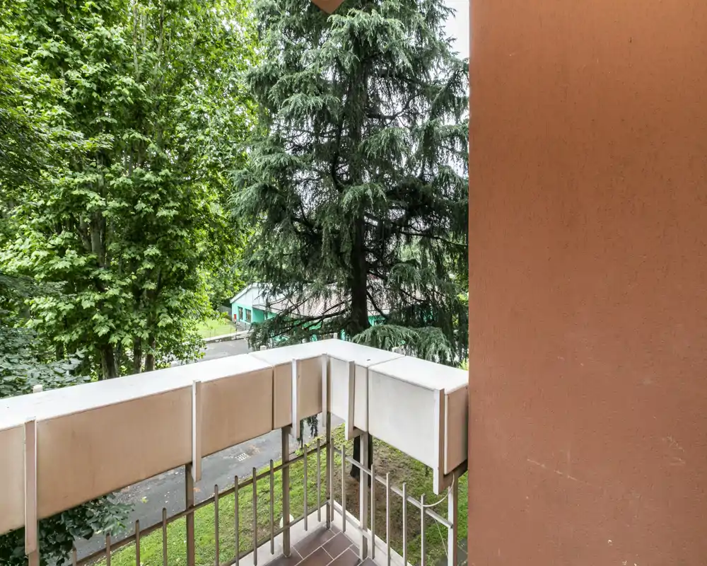Piccolo balcone privato con ringhiera in metallo e pavimento piastrellato, con vista su alberi maturi e una strada tranquilla — ideale per un caffè mattutino all'aperto.