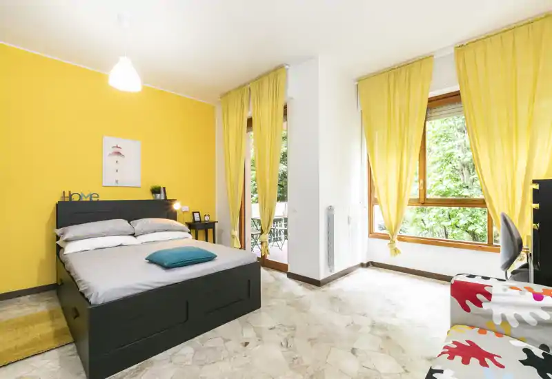 Camera da letto luminosa e di stile con letto in legno scuro, parete gialla e grandi finestre che lasciano entrare molta luce naturale. Biancheria confortevole, lampade da comodino e un piccolo balcone rendono lo spazio accogliente e pronto per l'affitto.