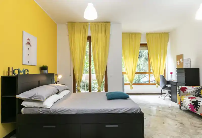 Ampia e luminosa camera da letto con un grande letto matrimoniale, parete d'accento gialla e ampie finestre che illuminano l'ambiente. Angolo studio e poltrona colorata rendono lo spazio versatile e accogliente.