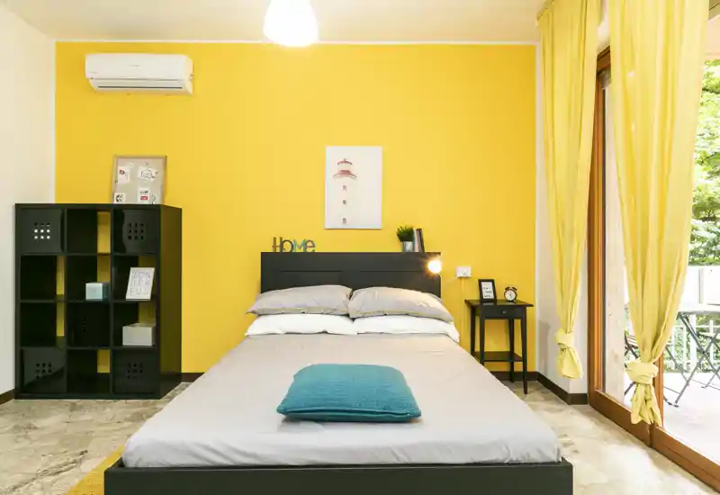 Camera da letto moderna e luminosa con letto matrimoniale centrato su una parete gialla, comodino, scaffalatura e porte-finestre che si aprono su un balcone.
