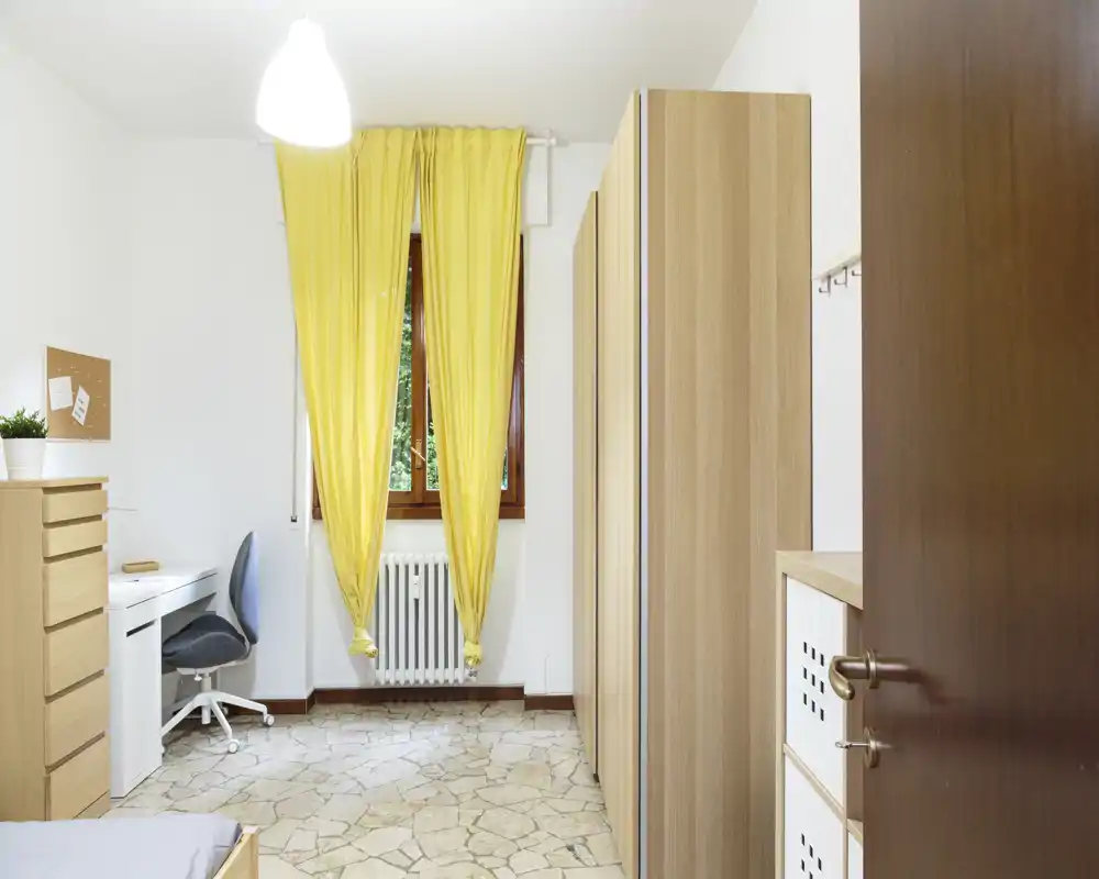 Camera da letto compatta con angolo studio: letto parzialmente visibile, scrivania bianca e sedia, armadi alti in legno e tende gialle che lasciano entrare luce naturale.