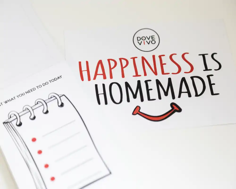 Primo piano di carte stampate su una superficie bianca con la scritta "Happiness is Homemade" e un'illustrazione di un bloc notes; nessun contesto della stanza visibile.