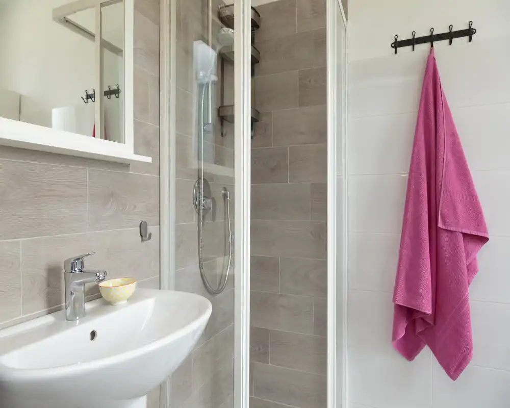 Bright, modern bathroom with a glass shower enclosure, wall-mounted sink and a pink towel on wall hooks. Clean neutral tiles and contemporary fixtures create a fresh, functional space.