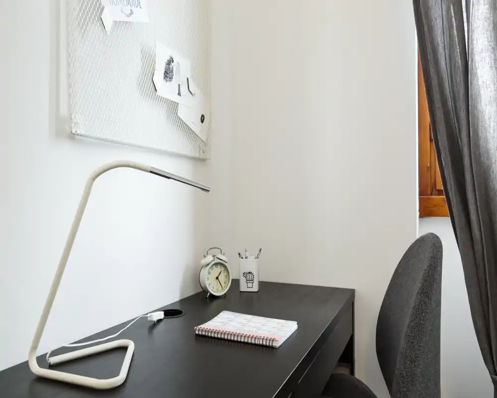 Compact workspace with a modern desk lamp, alarm clock and notebook beside a comfortable upholstered chair next to a curtained window — ideal for studying or remote work.