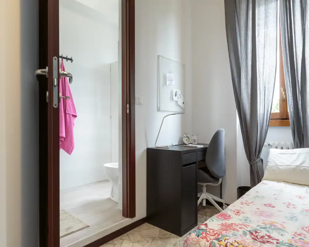 Bright small bedroom with a floral bedspread, a compact workspace with a desk and chair, and an en-suite bathroom visible through a wooden-framed door.
