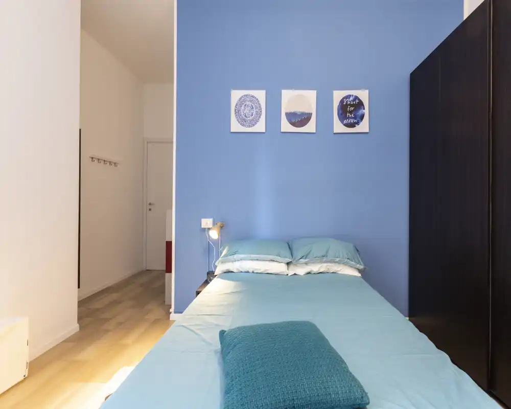 Camera da letto moderna e luminosa con letto matrimoniale ben rifatto su una parete blu, cuscini morbidi e un plaid che rendono l'ambiente accogliente.
