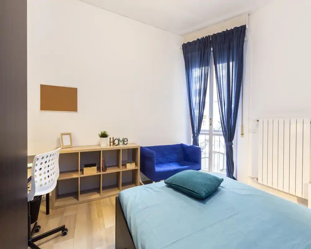 Camera da letto luminosa e moderna con letto ben rifatto in primo piano, un piccolo divano blu vicino alla finestra, una scrivania con scaffalatura e luce naturale da una finestra alta con tende blu scuro.