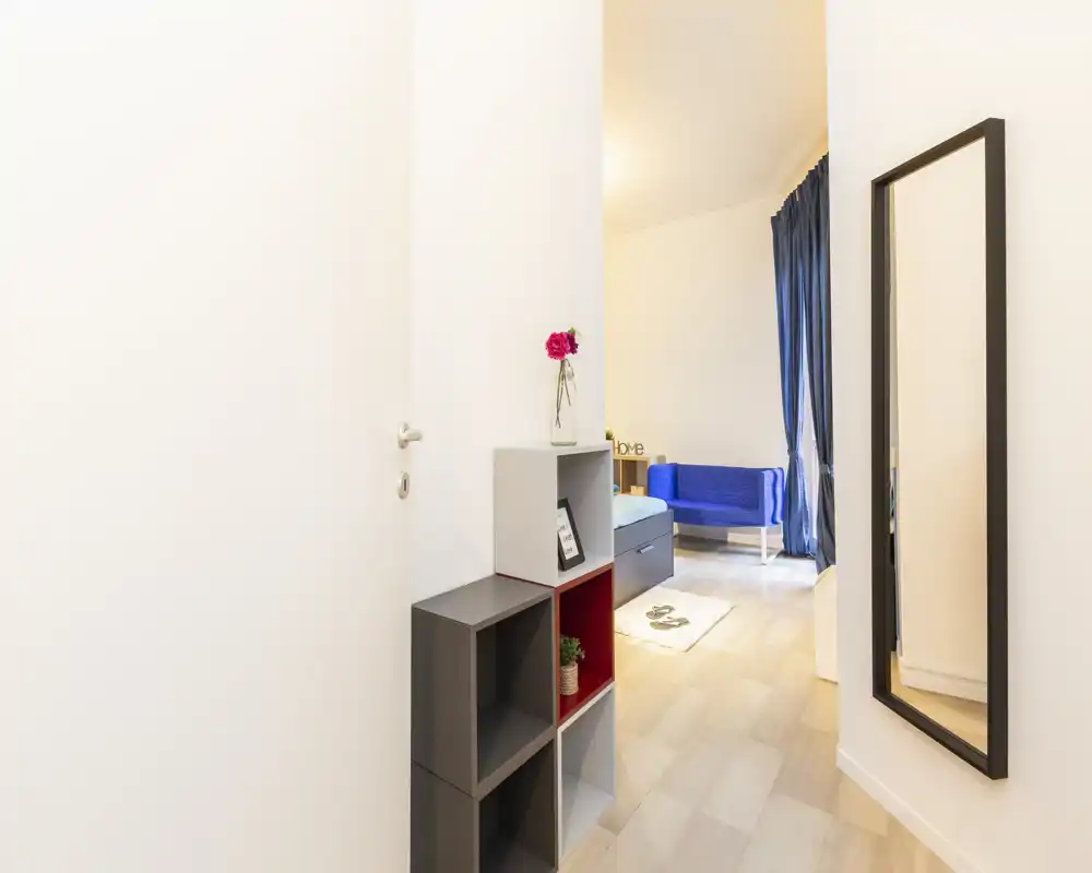 Piccolo corridoio d'ingresso ben illuminato con scaffalatura a cubi moderna e uno specchio a figura intera, che conduce a una zona giorno con divano blu e tende.