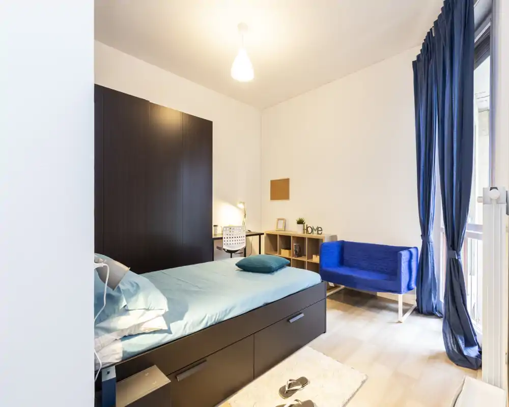 Camera da letto luminosa e moderna con letto singolo, cassettoni incorporati, una piccola scrivania e un divano blu vicino alla finestra — ideale per studenti o single.