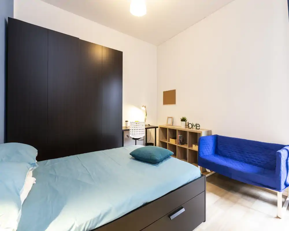 Camera da letto moderna e luminosa con letto matrimoniale rifatto, scrivania compatta con lampada, scaffalatura e un piccolo divano blu — ben allestita per affitti.