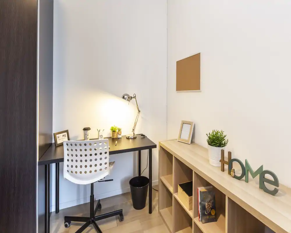 Angolo home office compatto con scrivania nera, lampada da lavoro e sedia girevole bianca, arricchito da piccole piante e scaffalatura per riporre oggetti — spazio di lavoro luminoso e ordinato ideale per il lavoro da remoto.