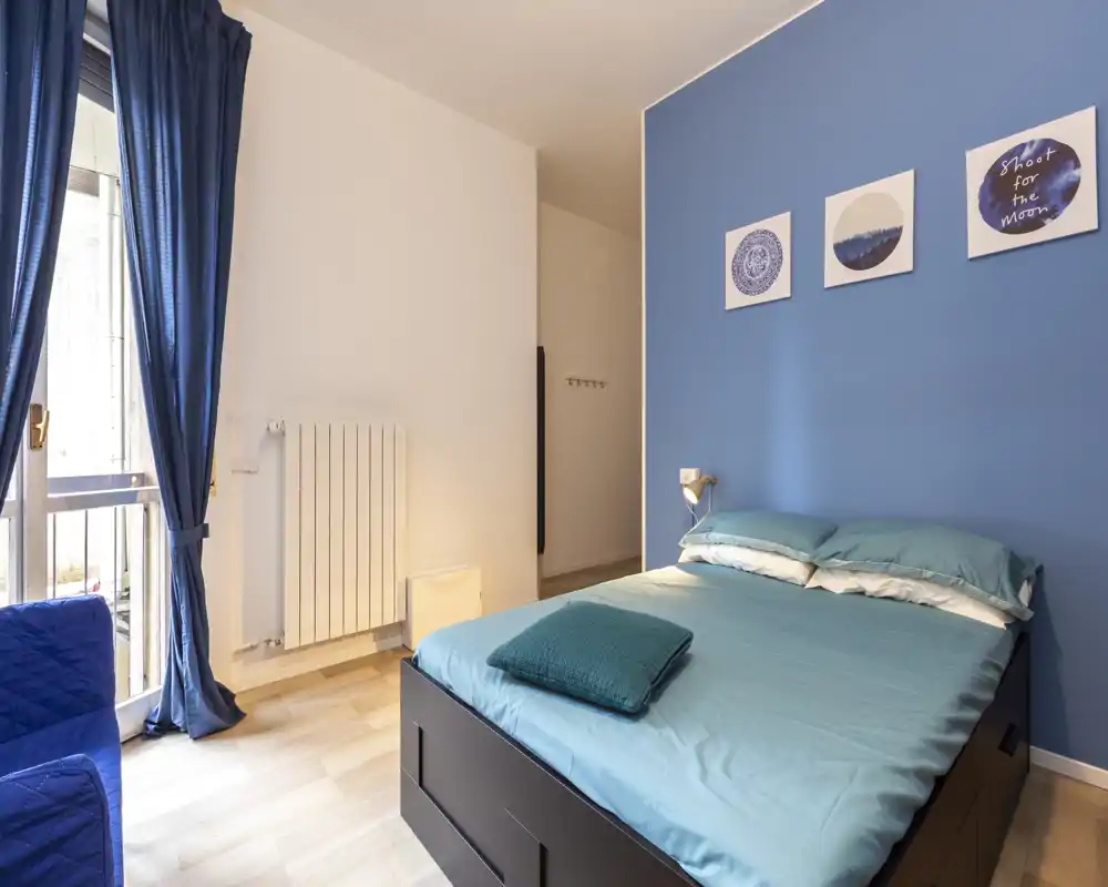 Camera da letto luminosa e ordinata con letto matrimoniale contro una parete blu, biancheria coordinata e luce naturale dalla finestra con tende.