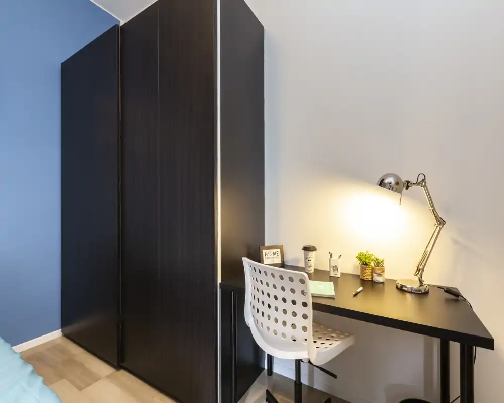 Angolo camera da letto luminoso e moderno con un armadio scuro e una scrivania ordinata illuminata da una lampada da tavolo; spazio studio compatto con sedia, piante e accessori.