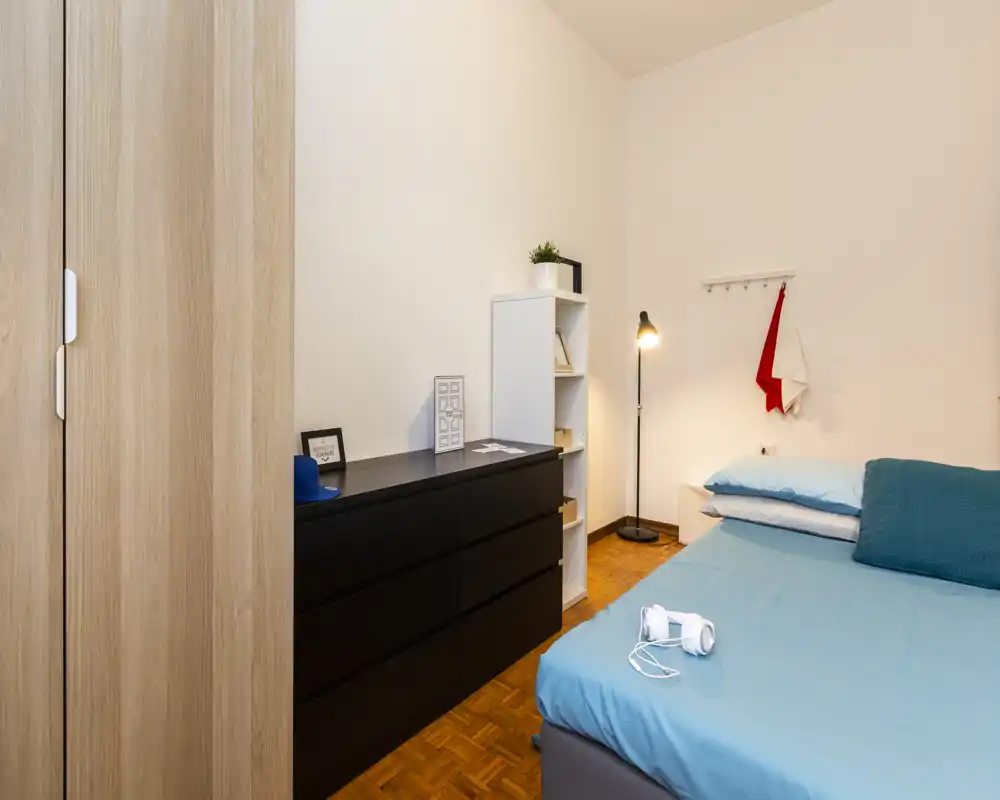 Bright, simply styled bedroom with a double bed dressed in blue linens, a large dresser and a standing lamp providing warm light — ideal for rental listings highlighting a tidy private room.