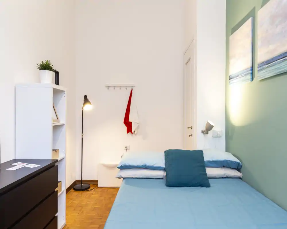 Neat, compact bedroom with a blue-dressed double bed, bedside reading lamp and simple wall art; bright, well-lit and tastefully styled for short-term rental listings.