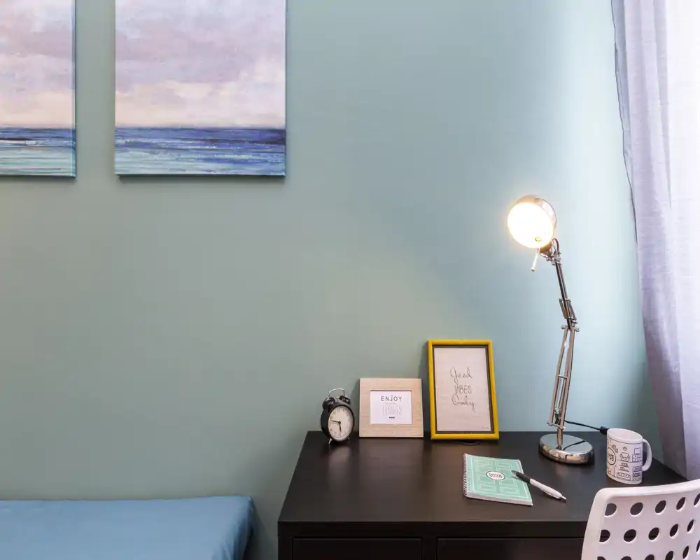 Compact bedroom corner with a dark wood desk, task lamp, framed prints and a portion of a blue bed visible at the left — clean, bright workspace within the sleeping area.