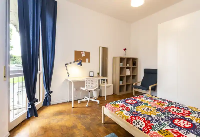 Bright, neatly styled bedroom with a single wooden bed dressed in colorful floral bedding, a study desk by the window, shelving and a comfortable armchair — ideal for a student or solo renter.