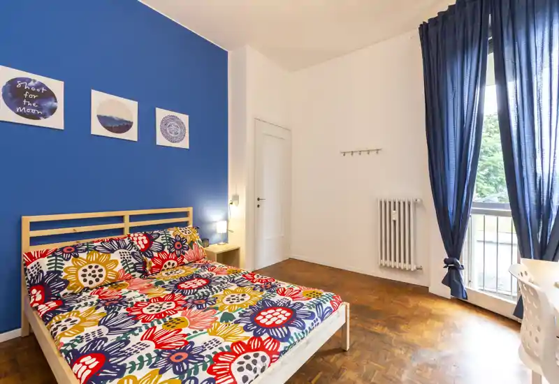Bright, stylish bedroom with a wooden bed dressed in a colorful floral duvet, a bold blue accent wall and large window with navy curtains providing abundant natural light.