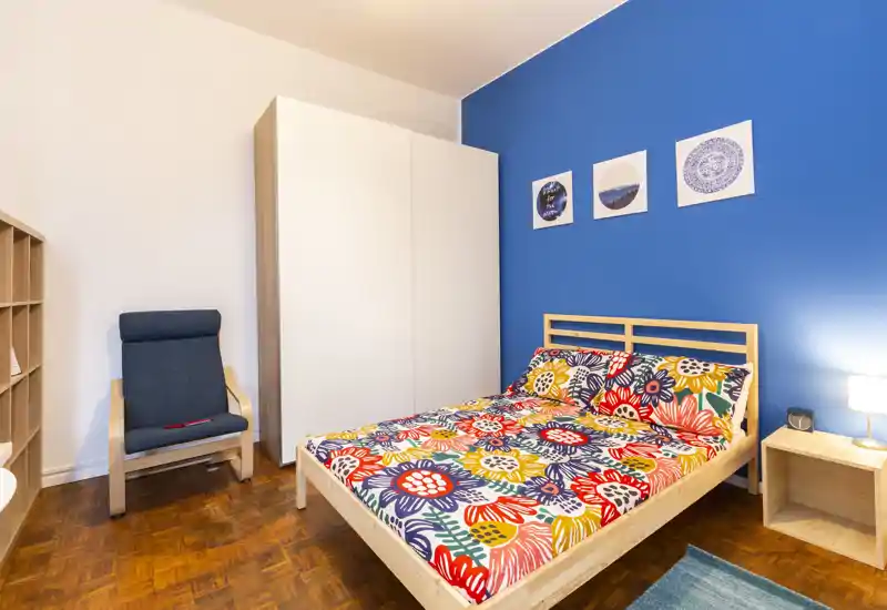 Bright, colorful bedroom with a wooden bed frame, patterned bedding and a blue accent wall. The room includes a wardrobe, bedside table with lamp and a comfortable armchair — ideal for a cozy single or double stay.