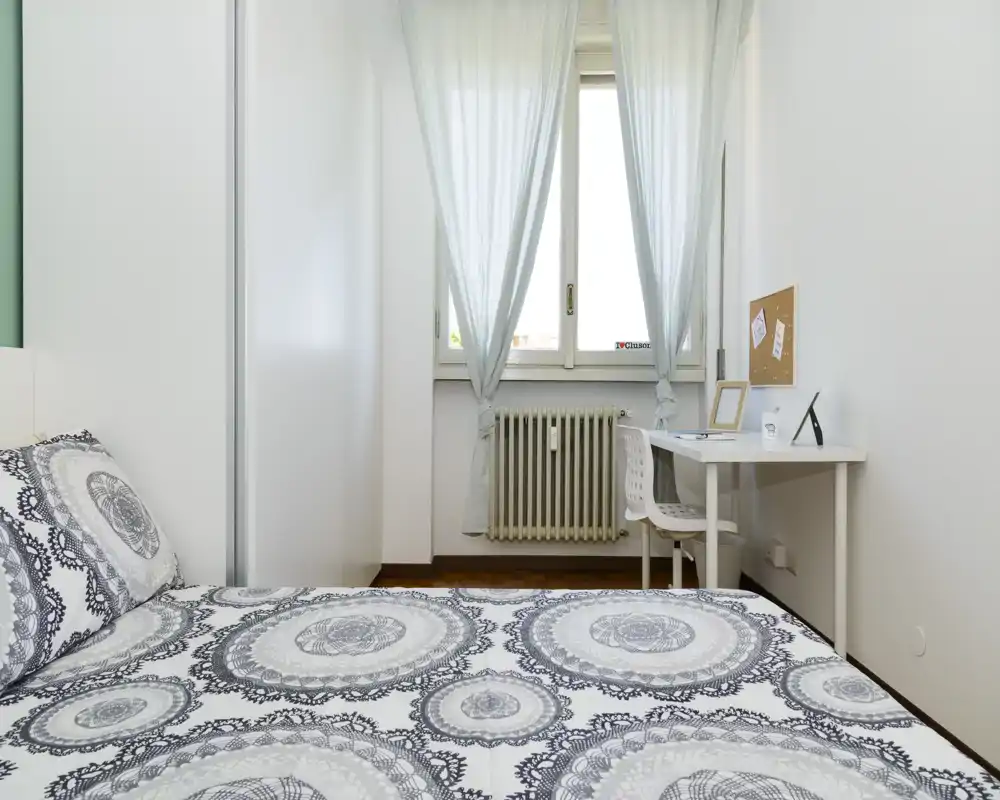 Camera da letto luminosa e minimalista con biancheria a motivo, scrivania vicino alla finestra e luce naturale soffusa.
