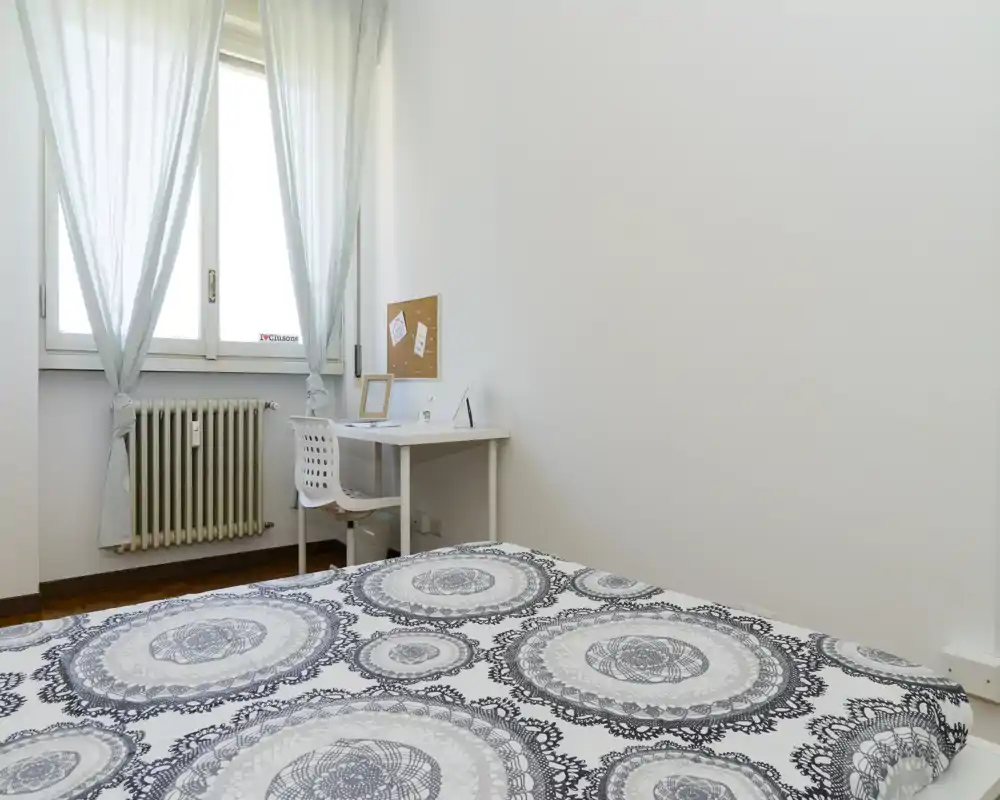 Camera da letto luminosa e minimal con un letto matrimoniale con copriletto decorato in primo piano e una piccola scrivania vicino alla finestra — ideale per chi cerca una stanza ordinata e ben illuminata.