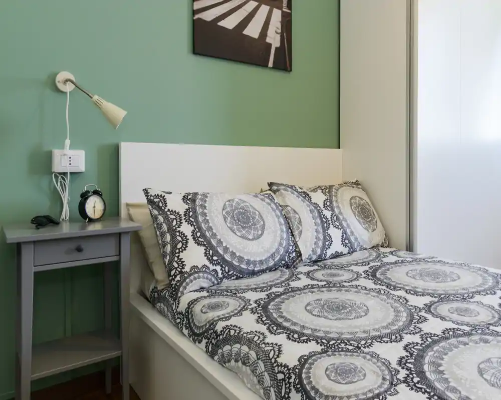 Camera da letto luminosa e ben arredata con copriletto a motivo e cuscini coordinati, comodino con lampada e sveglia — perfetta per annunci di affitto.