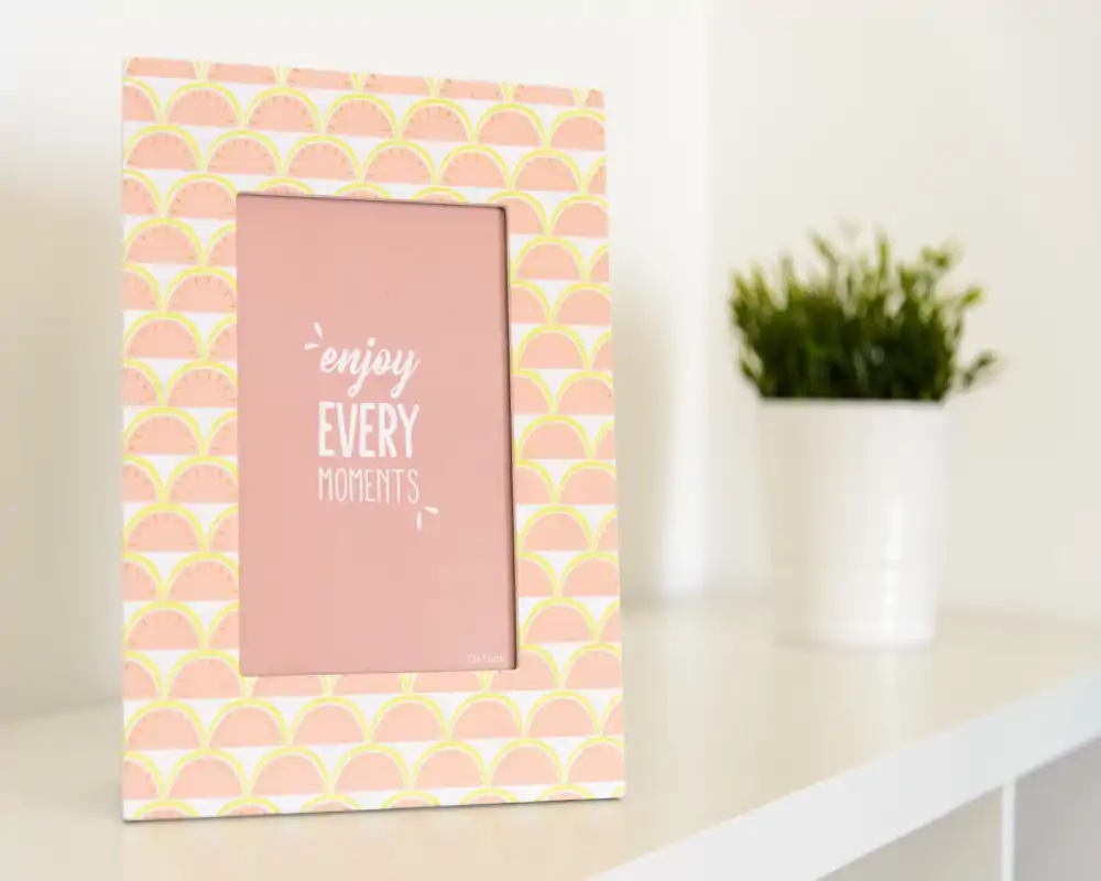 Primo piano di una cornice decorativa con la scritta "enjoy EVERY moments" su una mensola bianca, con una piccola pianta in vaso sullo sfondo; ideale come foto di dettaglio d'arredo.