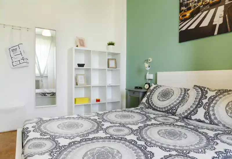 Camera da letto doppia luminosa e ben arredata con biancheria a motivo, comodino e scaffale; parete verde a contrasto e grande specchio che amplificano lo spazio.