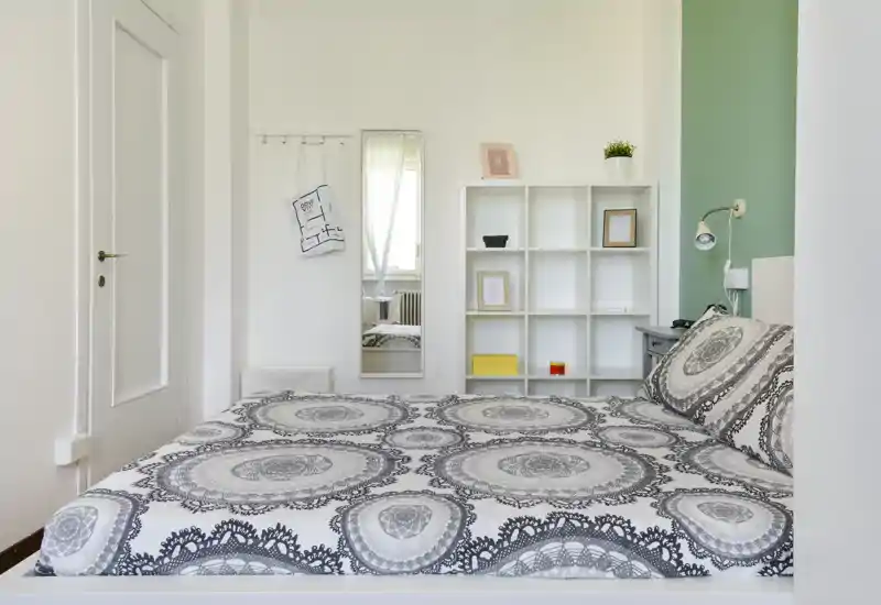 Camera da letto luminosa e moderna con un letto matrimoniale completamente visibile, biancheria a motivo, mensole bianche e una piccola lampada da comodino; pulita, ben illuminata e arredata in modo semplice per foto di affitti.