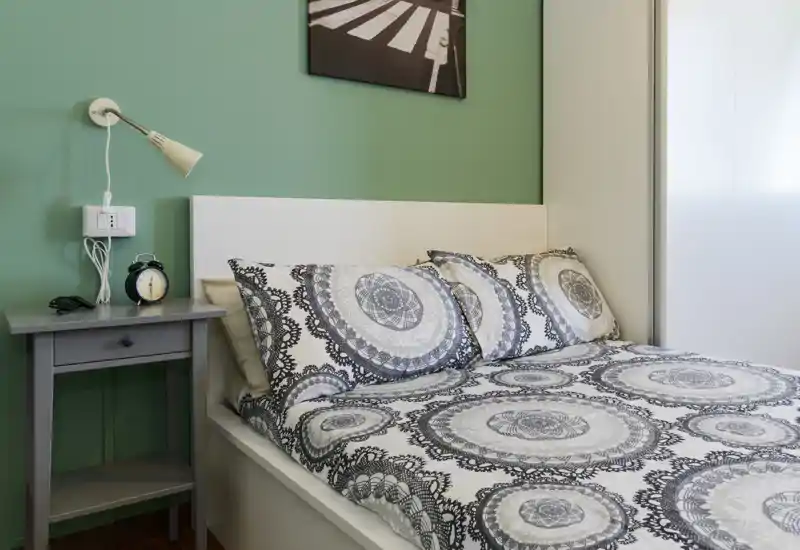 Camera da letto luminosa e ben arredata con copriletto a motivo e cuscini coordinati, comodino con lampada e sveglia — perfetta per annunci di affitto.