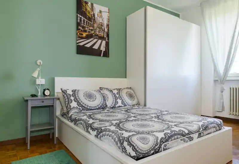Camera da letto luminosa e ordinata con letto matrimoniale, biancheria a motivi, comodino e armadio. La luce naturale e la parete verde creano un’atmosfera accogliente.
