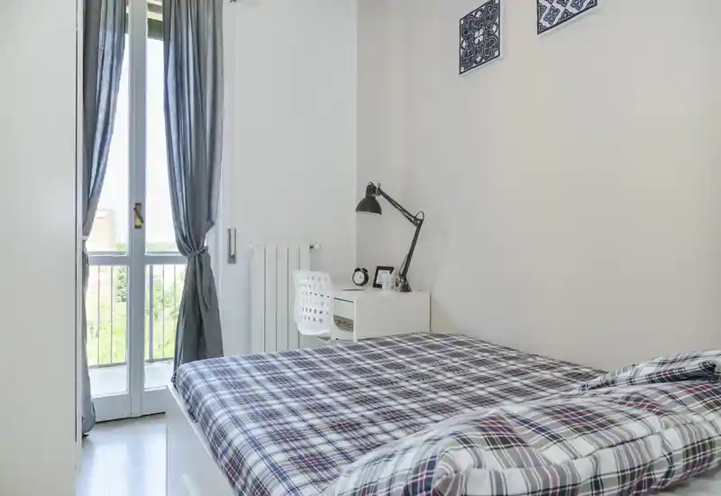 Bright, neatly styled bedroom with a double bed dressed in plaid bedding, a small desk with lamp and chair by a balcony window that fills the room with natural light.